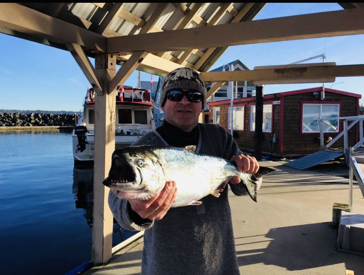 Douglas presenting a fish he caught to the camera on a dock. Calm waters, sunny skies and a small building are visible in the background.